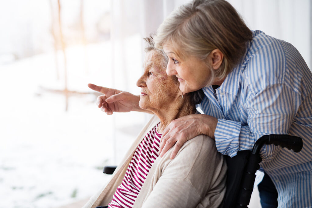 An elderly mother with her daughter at home looking out of the window. A caregiver assisting a disabled senior woman in wheelchair. November is National Family Caregiving Month