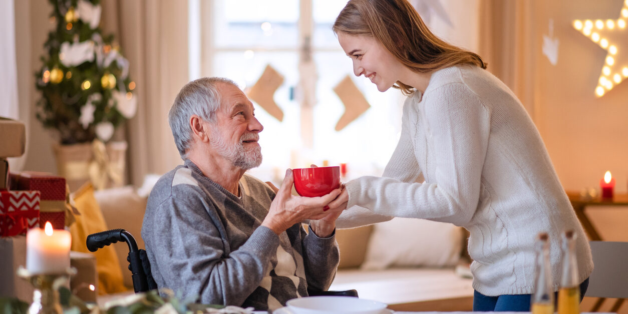 Young woman giving cup of tea to senior grandfather in wheelchair indoors at home at Christmas. Appreciating Family Caregivers During the Holiday Season