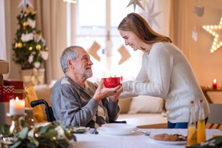 Young woman giving cup of tea to senior grandfather in wheelchair indoors at home at Christmas. Appreciating Family Caregivers During the Holiday Season